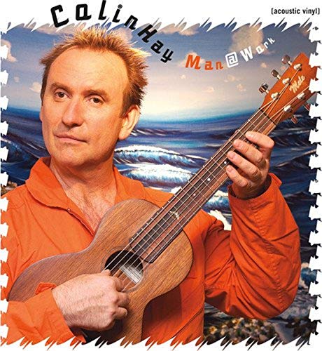 Colin Hay wearing an orange shirt and playing a wooden ukulele against ocean backdrop.