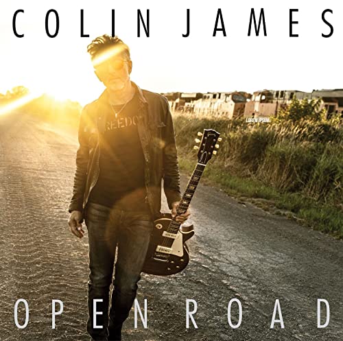 Colin James holding a guitar on a rural road at sunset.