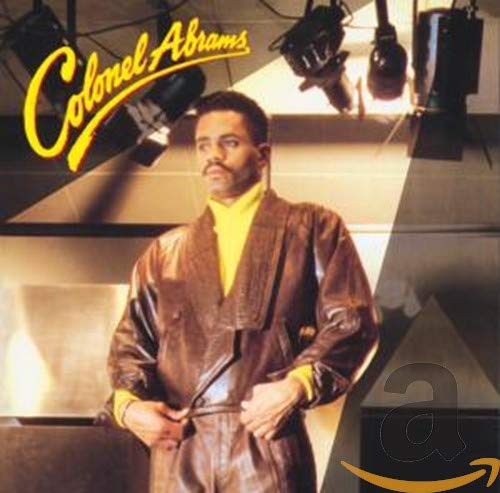 Colonel Abrams wearing a brown leather jacket and yellow shirt under studio lights.