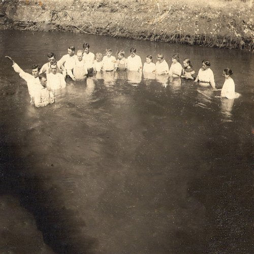 A sepia-toned photograph of a group baptism in a river, people wearing white robes.