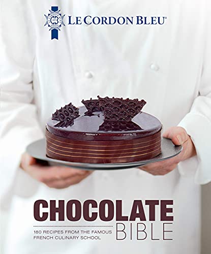 A chef in a white uniform holds a chocolate cake on a plate.