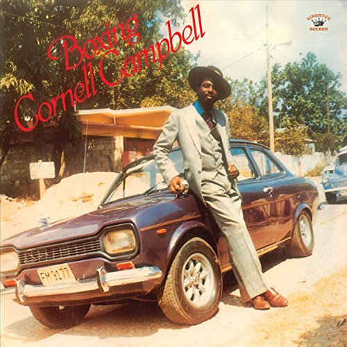 Cornell Campbell in a grey suit leaning against a purple car on a dirt road.
