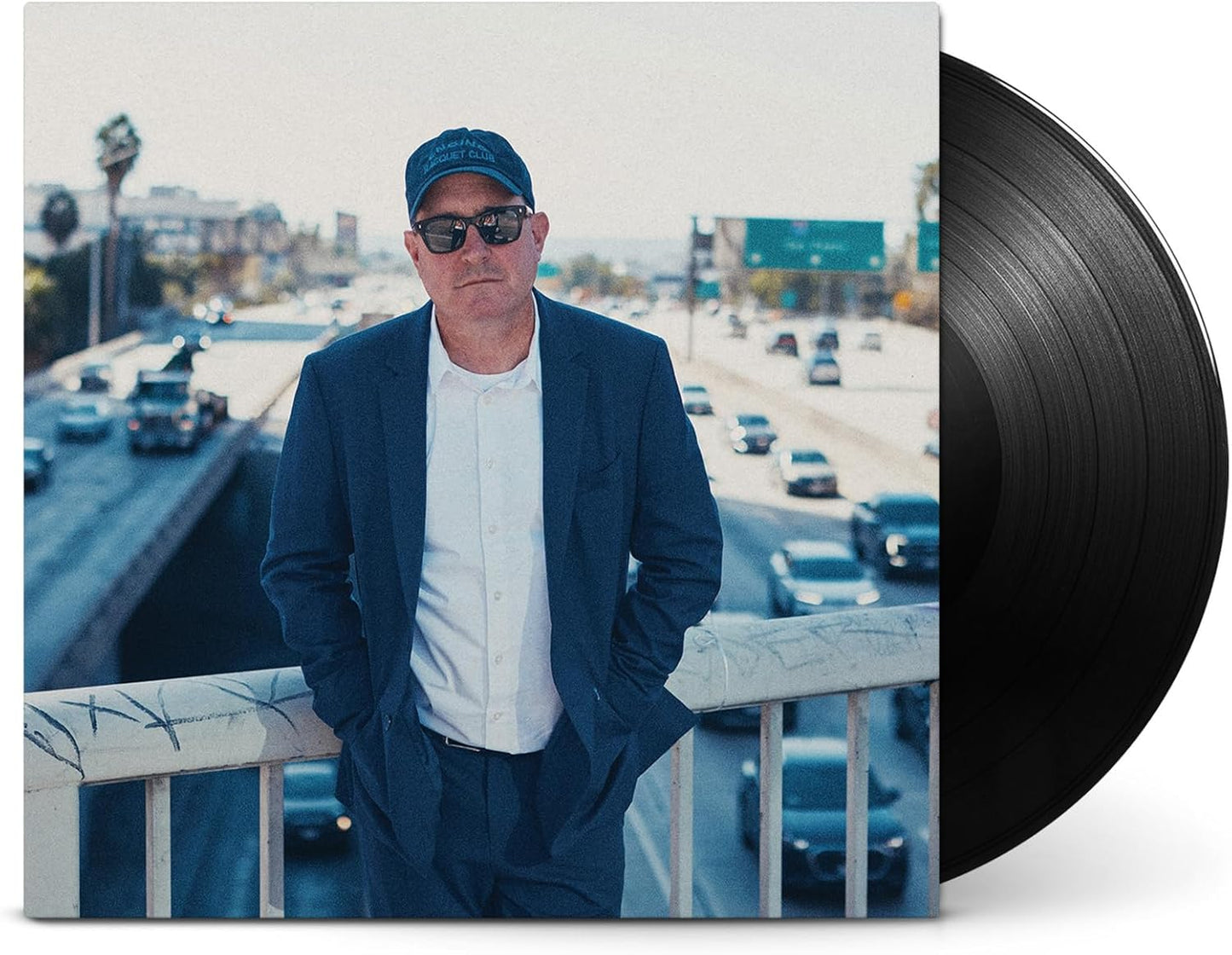 Craig Finn in a suit and cap standing on a bridge over a highway.