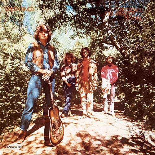 Four men standing on a wooded path, one holding a guitar.