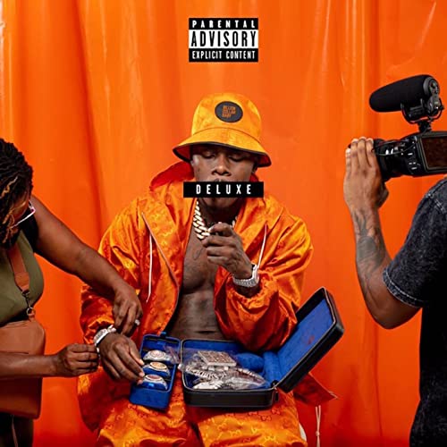 DaBaby wearing an orange bucket hat and jacket, holding jewelry before an orange backdrop.