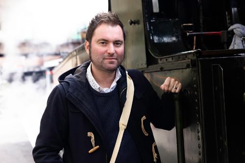 Man in coat stands near a vintage steam train engine.