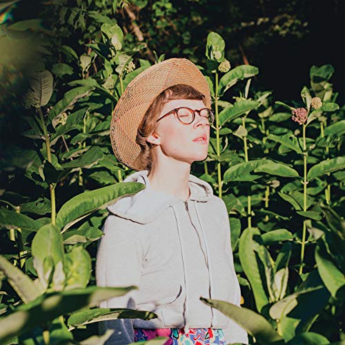 The image depicts a woman wearing a straw hat and glasses, surrounded by tall plants. Alt text: Woman in a straw hat standing among tall green plants.