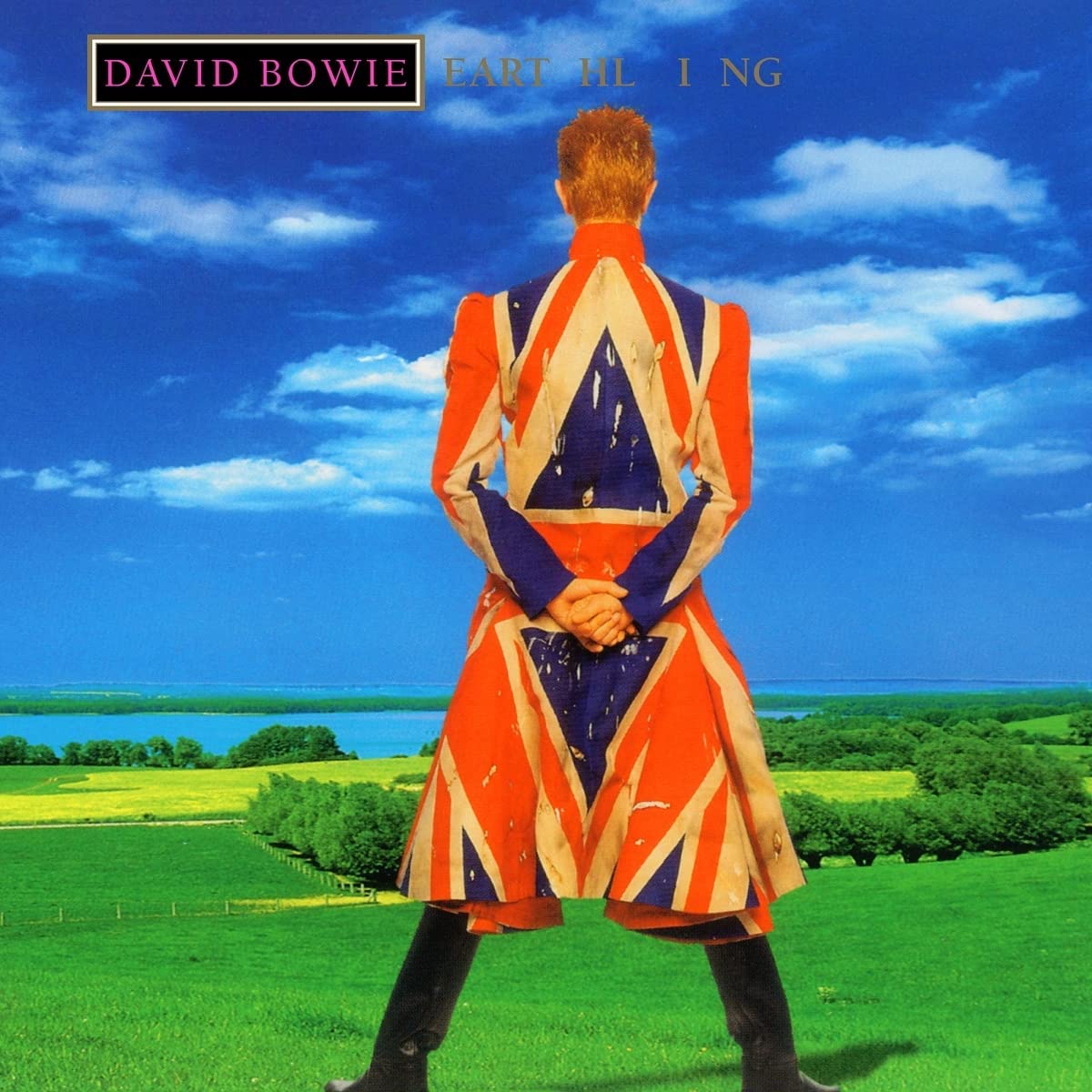 David Bowie stands in a field wearing a Union Jack coat, facing away from camera.