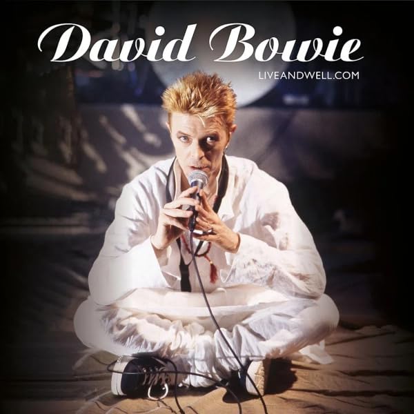 David Bowie sitting cross-legged on the floor holding a microphone, wearing white clothing.