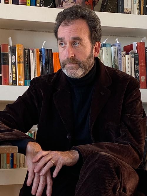 Man with beard in black jacket, bookshelves in background.