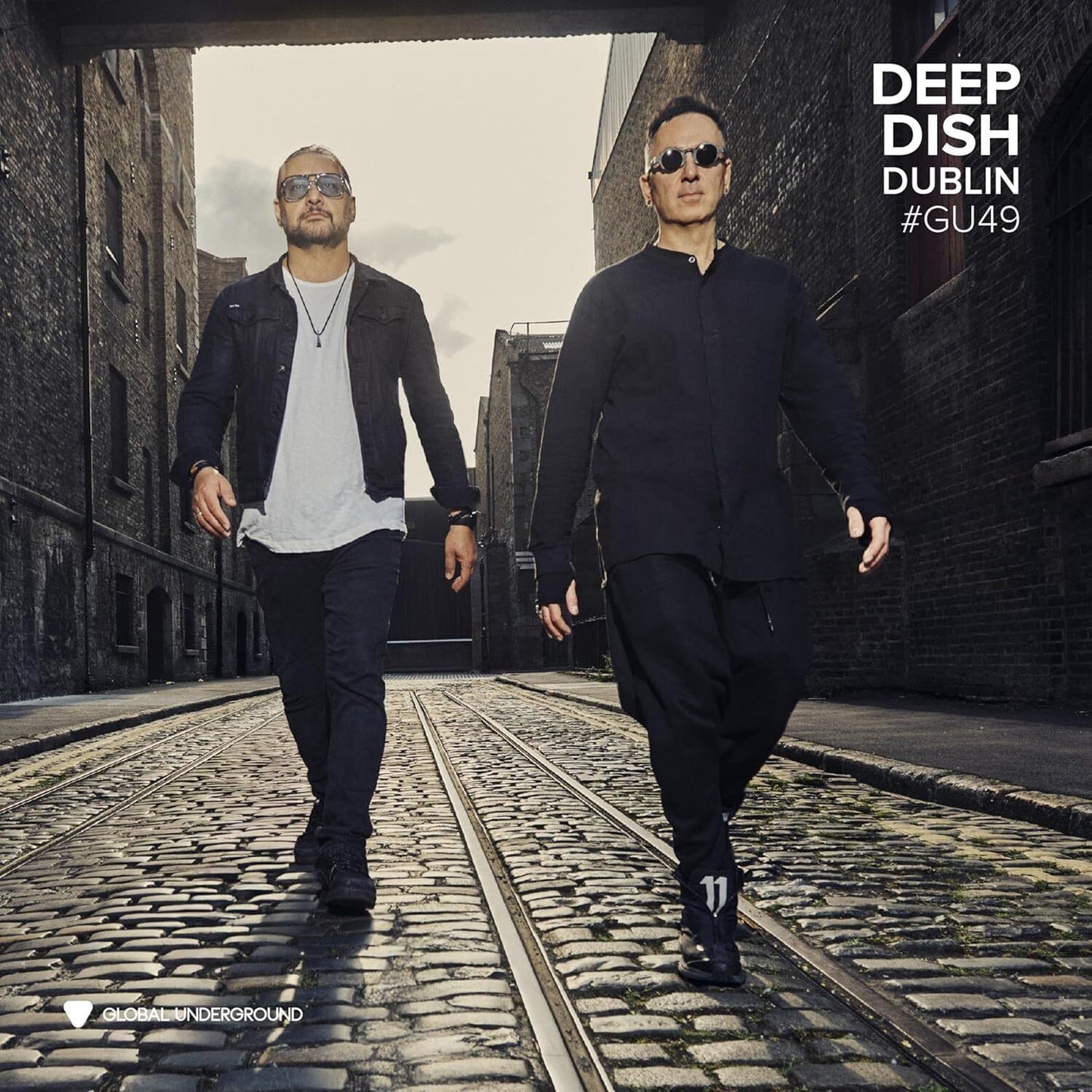 Two men in sunglasses walk along a cobblestone street between brick buildings.