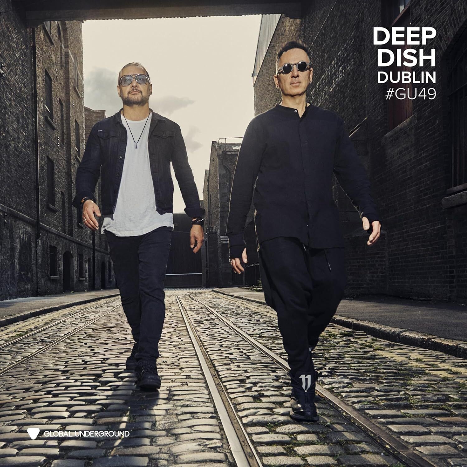 Two men in sunglasses walk along a cobblestone street between brick buildings.