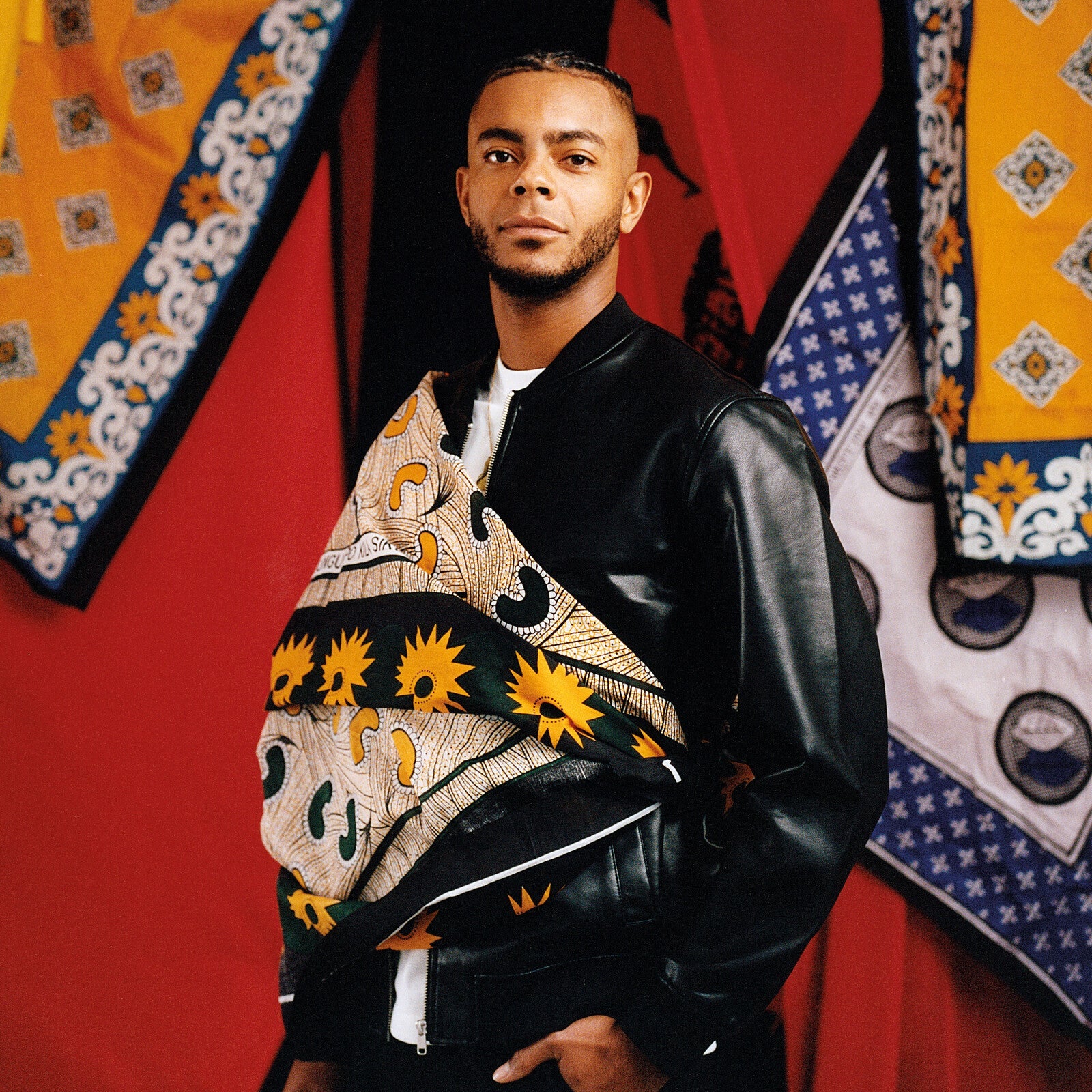Man in a patterned jacket stands before colorful textile backdrops.