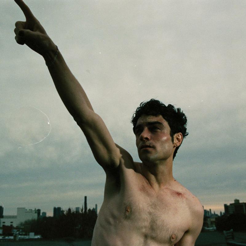 Shirtless man raising arm, city skyline blurred in background.
