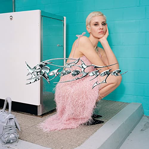 Blonde woman in pink dress seated against a teal wall.