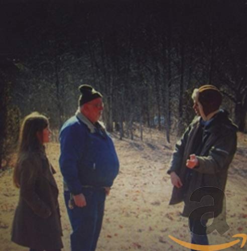 Three people stand in a wooded area, facing each other.