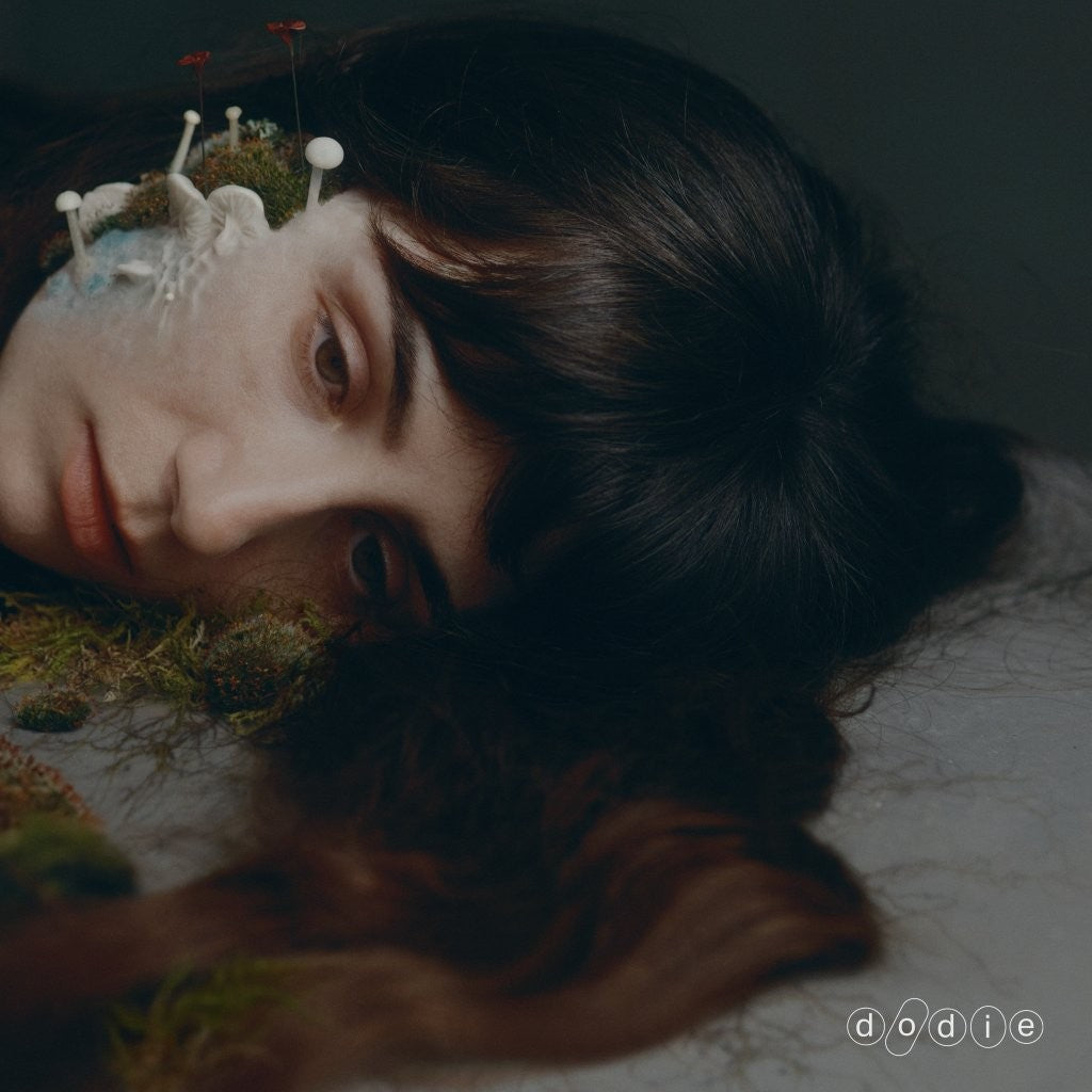A person with dark hair lying down, with moss and mushrooms growing on their face.