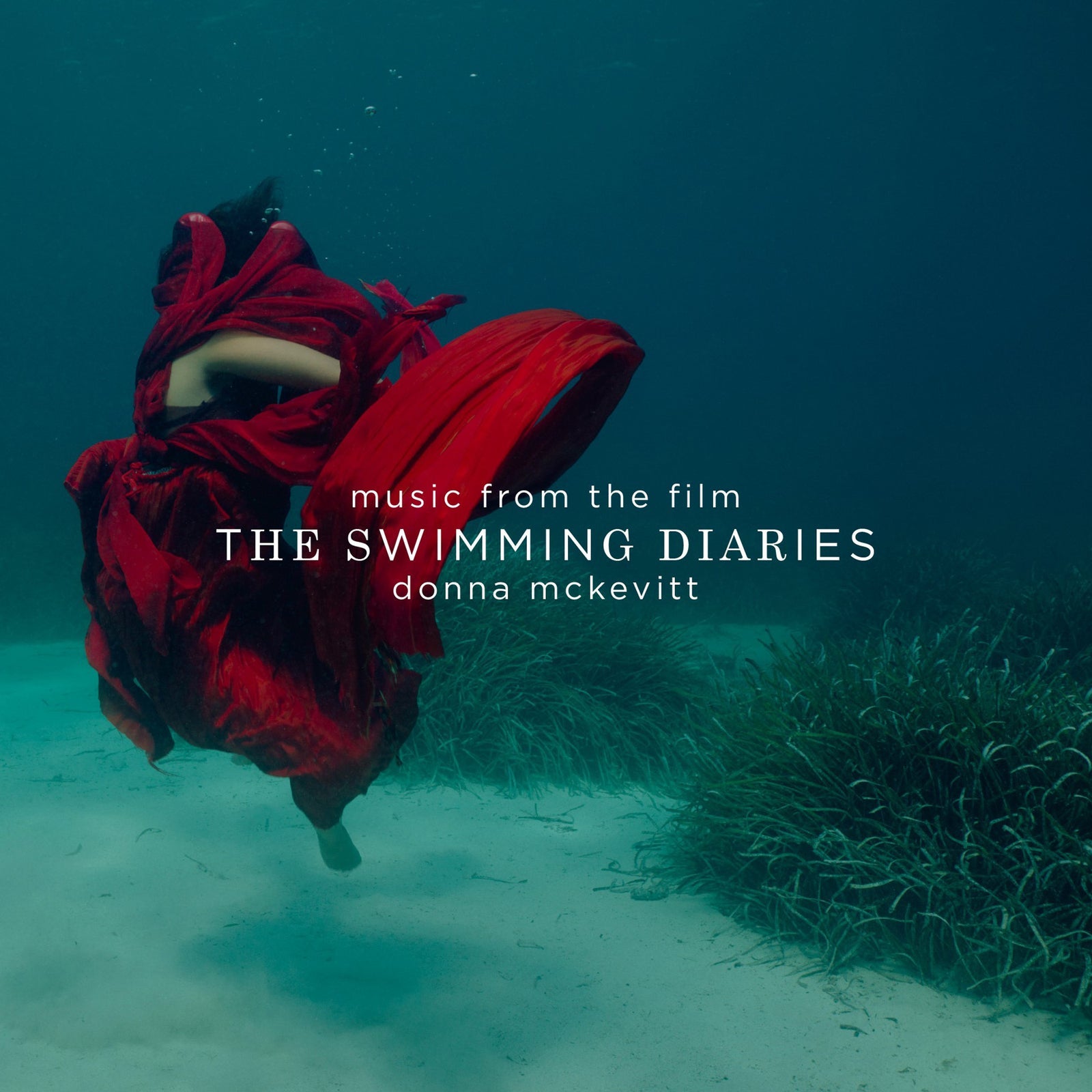 A person wrapped in flowing red fabric floating underwater above a sandy seabed.