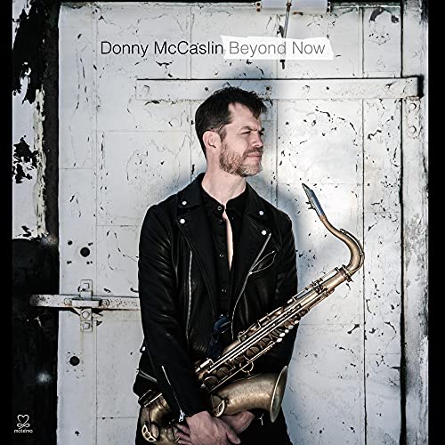 Donny McCaslin in a black leather jacket holding a saxophone against a white door.