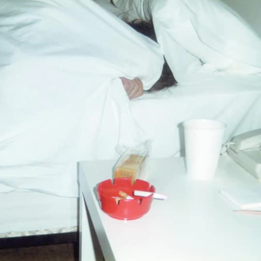 Person under white sheets with a bedside table and ashtray.