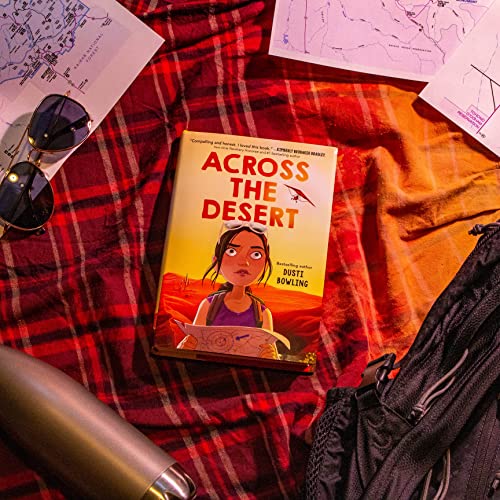 Book cover, "Across the Desert," with travel accessories on a red plaid blanket.