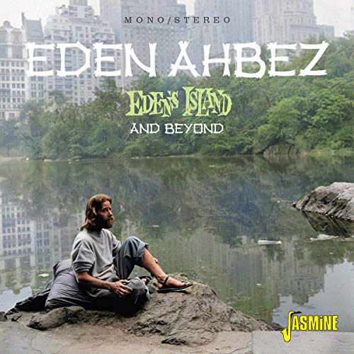 Eden Ahbez sits on a rock by a lake with city buildings in the background.