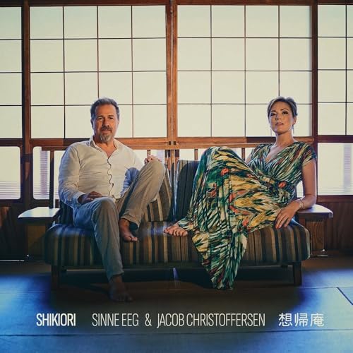 Album cover featuring two people seated on a striped sofa indoors.
