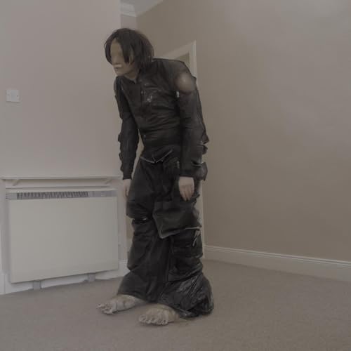 Person in dark, oversized clothing and a mask stands indoors.