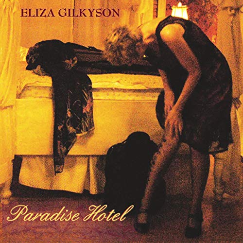 A woman in a dark dress stands in a dimly lit, amber-toned hotel room.