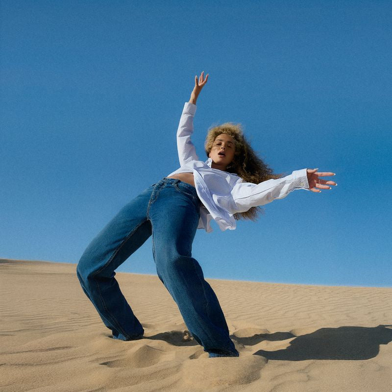 Ella Eyre in a white shirt and jeans posing in a sandy desert landscape.