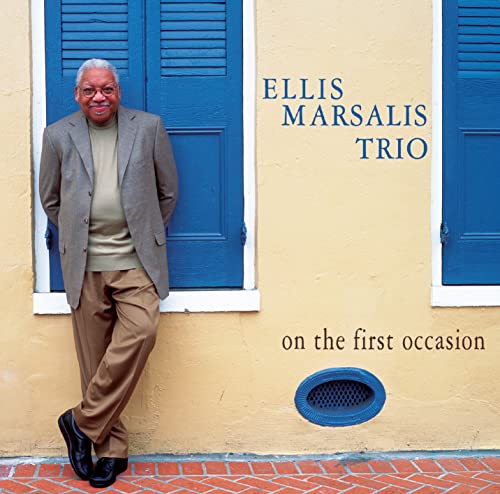 Ellis Marsalis poses in front of a yellow wall with blue shutters.