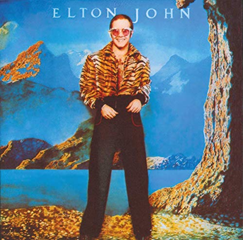Elton John in a tiger-print jacket, standing before a mountain landscape.