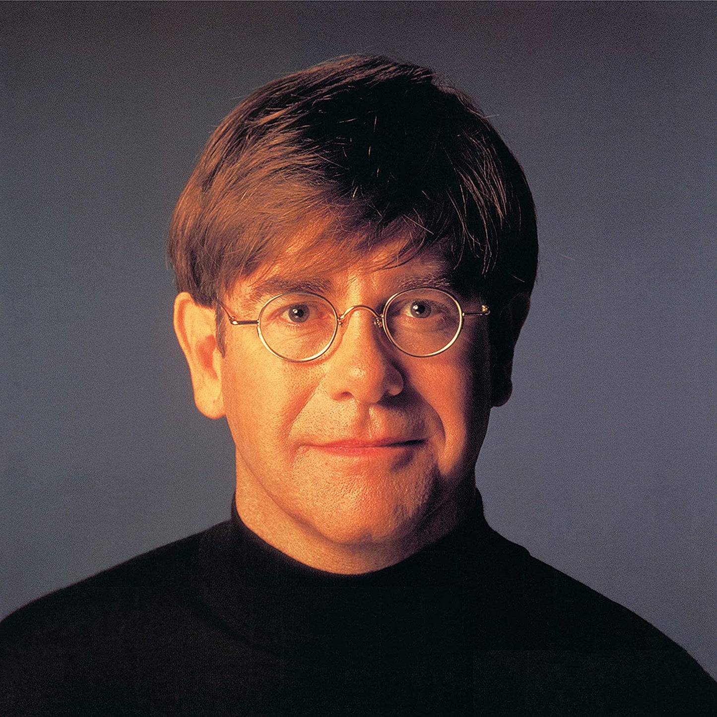 Elton John wearing glasses and a black shirt, against a gray background.