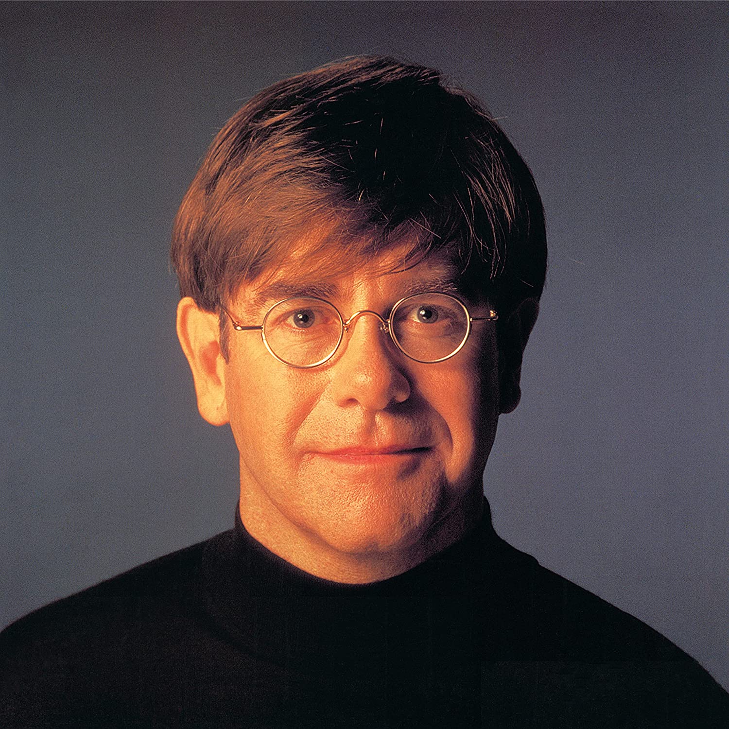 Elton John wearing glasses and a black shirt, against a gray background.