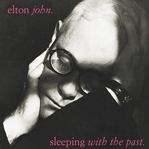 Black and white portrait of Elton John wearing glasses with hands on his head.