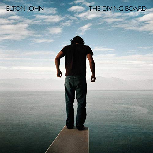 Man stands on diving board overlooking large body of water with mountains in distance.