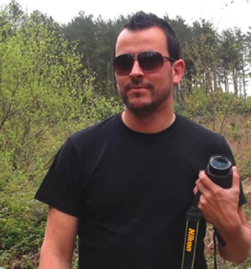 A man in a black t-shirt and sunglasses holding a Nikon camera outdoors.