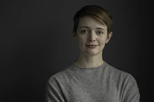 A person with short brown hair wearing a grey sweater against a dark background.