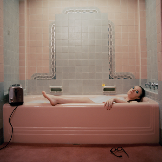 Woman in a pink bathtub with a toaster, pink tiled bathroom.
