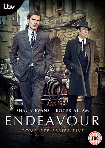 Two men in suits stand in front of a vintage car and stone building.