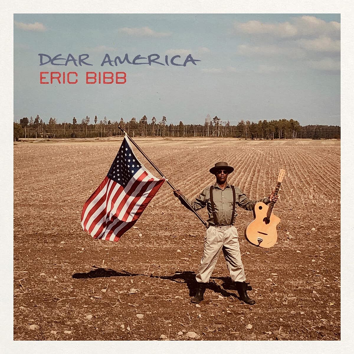 Eric Bibb standing in a field holding an American flag and an acoustic guitar.