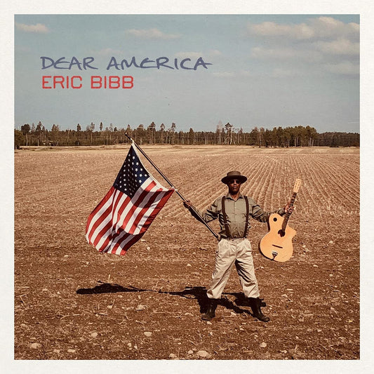 Eric Bibb standing in a field holding an American flag and an acoustic guitar.