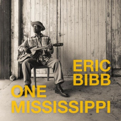 Eric Bibb sitting on a chair playing guitar in front of a wooden door.