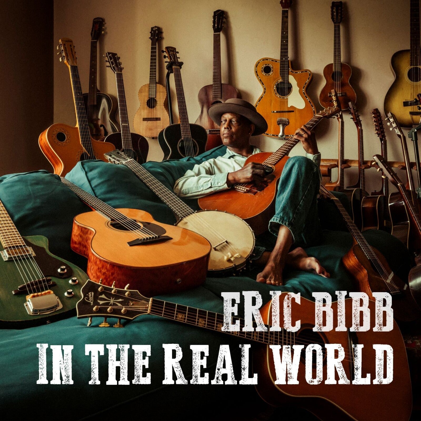Eric Bibb poses with guitars, album title “In The Real World” visible.