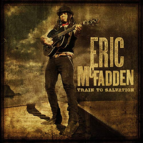 Eric McFadden wearing a hat and dreadlocks, playing an acoustic guitar on a landscape.