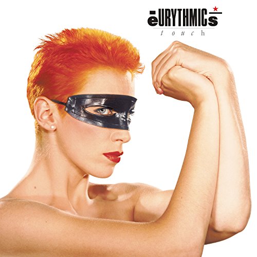 Annie Lennox with short orange hair wearing a black eye mask, posing against white.