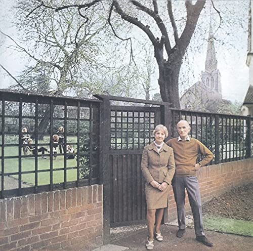 An older couple stands before a garden gate with a church in the background.