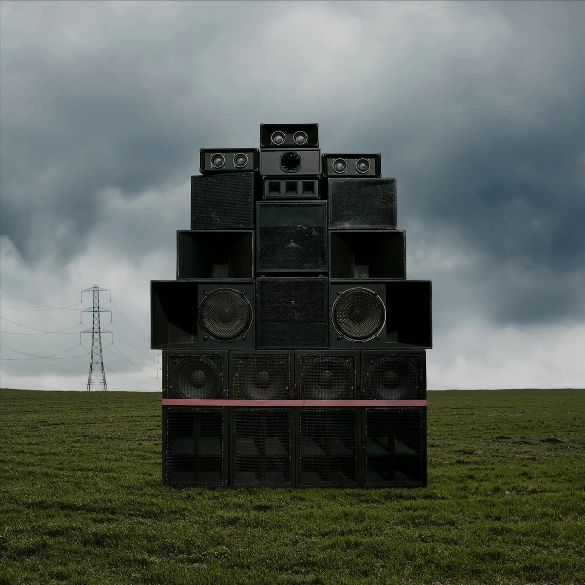A large stack of black speakers stands in a grassy field under cloudy skies.