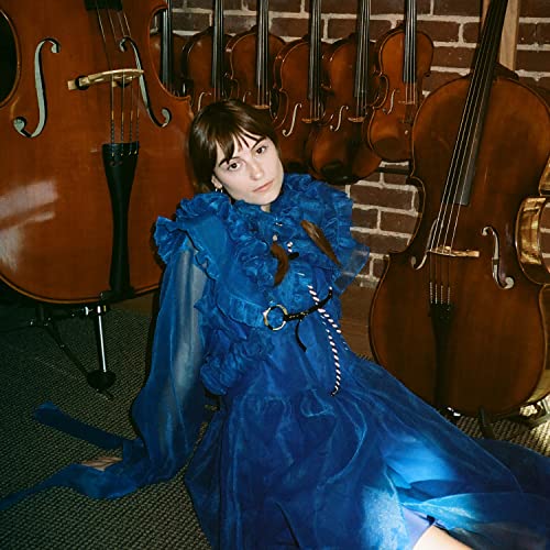 Faye Webster sits among several dark wood stringed instruments.