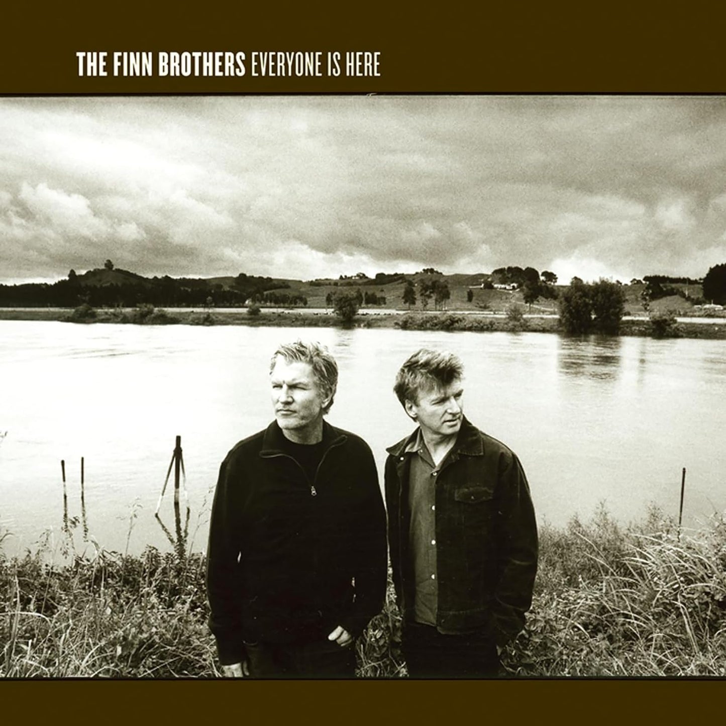 Two men stand by a river under a cloudy sky; album cover.